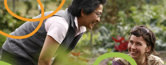 Two people standing in a vegetable garden, talking and smiling at one another.