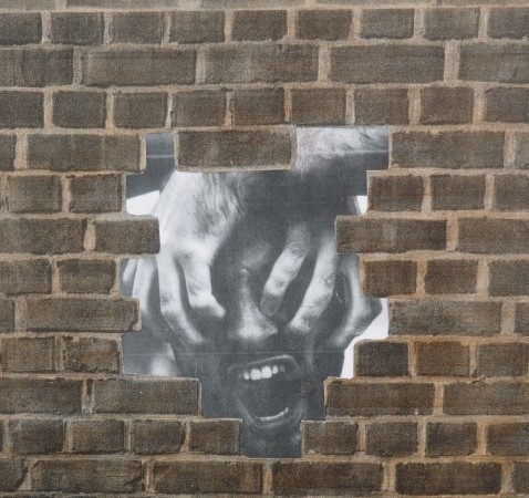 A brown brick wall, with a hole in the centre revealing a black and white drawing of a screaming face covered by two hands